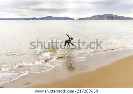 SAN FRANCISCO,USA - MARCH 1 2014: View of a surfer in wet suit surfing his board or skimboarding in shallow water in the Bay beach coast, sailboats crossing the sea in Marina,California,United States.