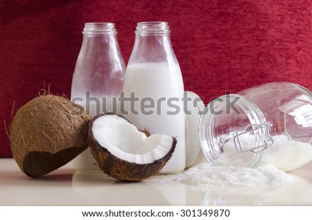 Coconut products. Cracked open coconut with meat cut in half, grounded flakes in a mason jar, flour and fresh milk in glass bottles on a table with red ruby background.