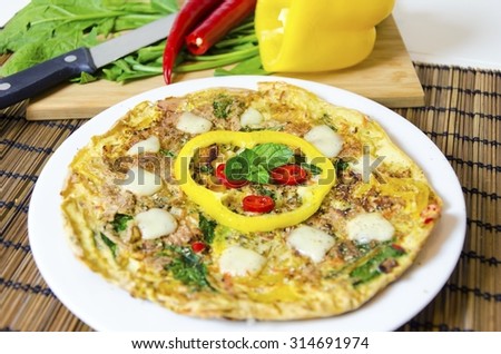 A spinach, chili, mint and pepper omelette in a dish over bamboo table cloth and chopping board with knife and raw vegetables on while background. A healthy diet meal omelet.