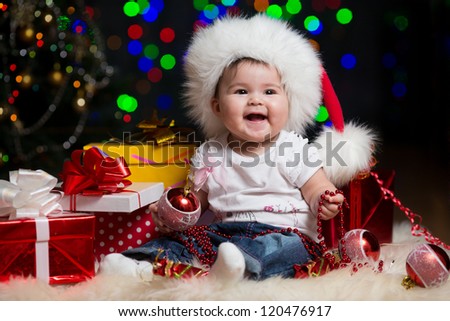 baby girl in Santa Claus hat with gifts under Christmas tree