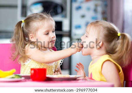 Two little children toddlers eating meal together, one girl feeding sister in sunny kitchen at home