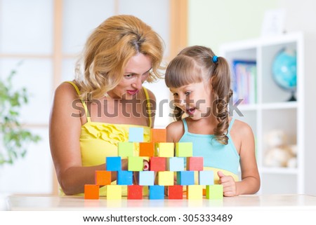 Middle aged mother helping her preschooler daughter build  tower from color building blocks as they sit together at table at home