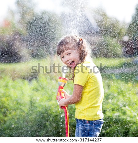 Cheerful kid girl watering plants from hose spray in garden at backyard of house at sunny summertime