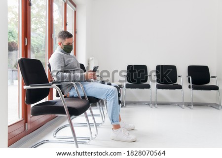 Young man with face mask sitting in a waiting room of a hospital or office looking at smartphone - focus on the man Foto stock © 
