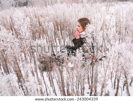 Beautiful young girl, sitting on the snow among frosty high grass. Breath warming cold hands.