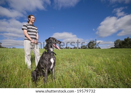 Male standing in meadow with dog on leash looking into horizon looking happy