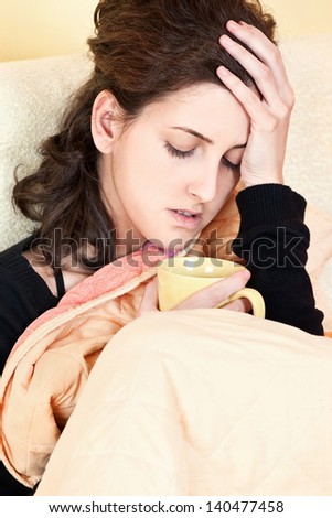 A young sick woman with a cup of tea covered with blanket at home, holding her head in pain