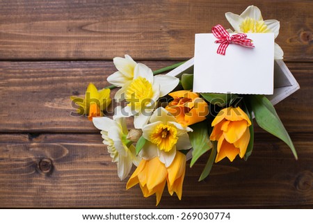 Fresh  spring yellow narcissus and  tulips flowers in wooden box and empty tag  on brown  painted wooden planks. Selective focus. Place for text.