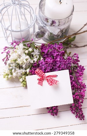 Postcard with fresh white and violet lilac flowers  and candles on white painted wooden planks. Selective focus. Place for text on empty tag.