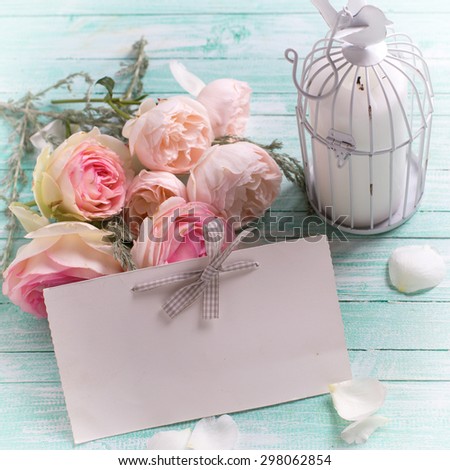 Background with fresh roses flowers, candle in decorative bird cage and empty tag on turquoise painted wooden background. Selective focus in on tag. Square image.