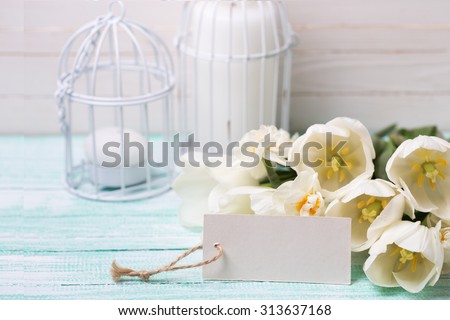 Postcard with white  flowers  tulips and daffodils and empty tag for your text on turquoise painted planks against white wall. Selective focus.