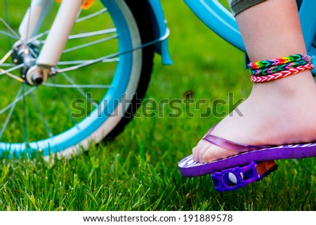 Young girl in flip-flops wearing loom bracelets on her hands and legs riding the bicycle. Close up. Young fashion concept