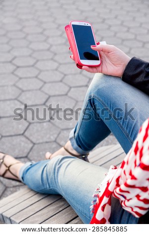Young woman holding a red covered smartphone. Sitting on a bench, wearing modern shoes, blue jeans, black jacket and a red white scarf. Fashion, technology, life style, online shopping, urban concept.