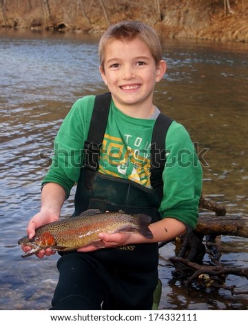 Boy fishing - Smiling youth in waders holding a Rainbow Trout fish next to a clear stream