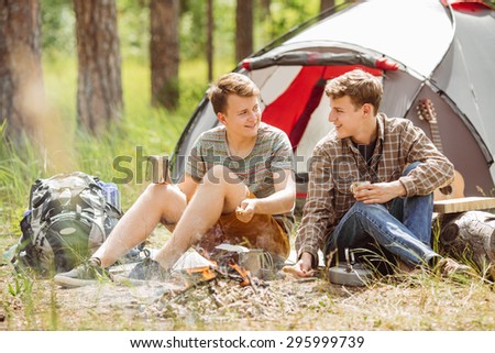 Two fellow campers talking to each other and drinking tea by a tent in the forest