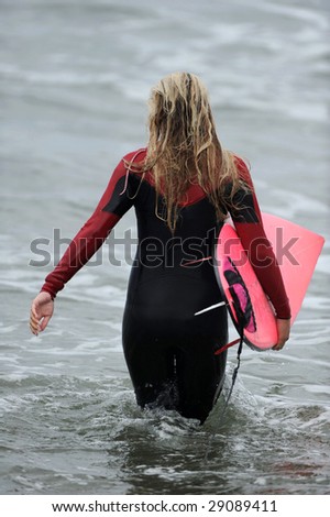 A female surfer with a pink board enters the ocean to go surfing.