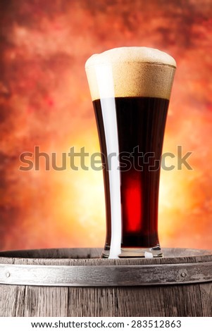 Tall glass of dark beer with foam and wooden barrel on orange background