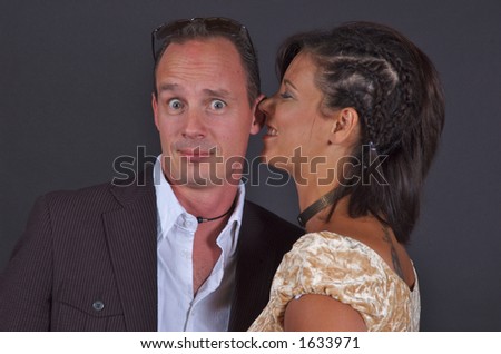 A woman whispering into a man\'s ear and smiling, and the man has a humorous expression on his face.