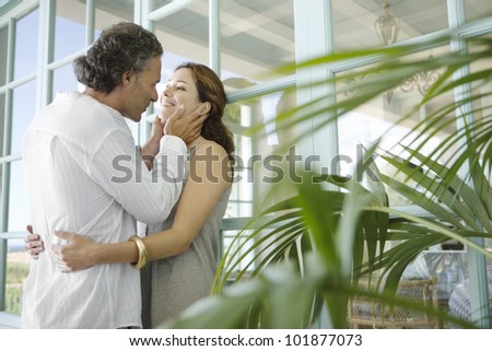 Mature couple hugging at home, standing by large garden doors and surrounded by foliage.