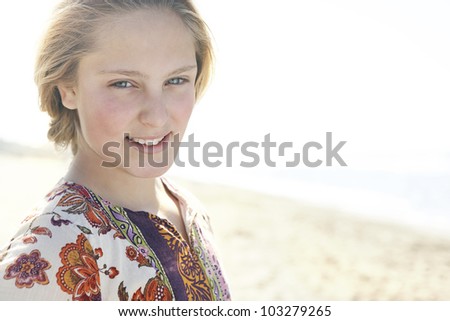 Close up of a blond girl standing on a beach shore, smiling at the camera.