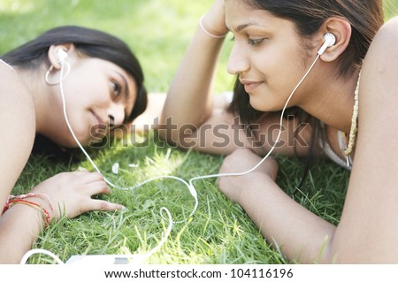 Two indian girls sharing their earphones to listen to music in the park.