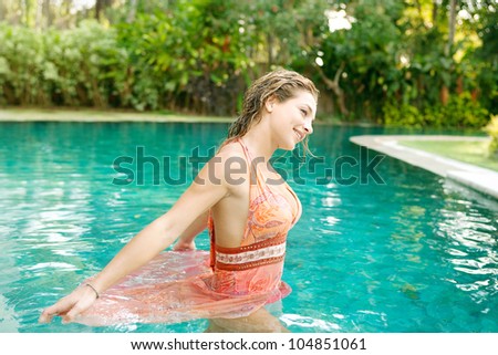 Portrait of a young woman in a swimming pool in a tropical garden, wearing a pink dress and smiling.