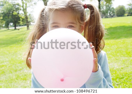 Young girl inflating a pink balloon in front of her face in the park on a sunny day.