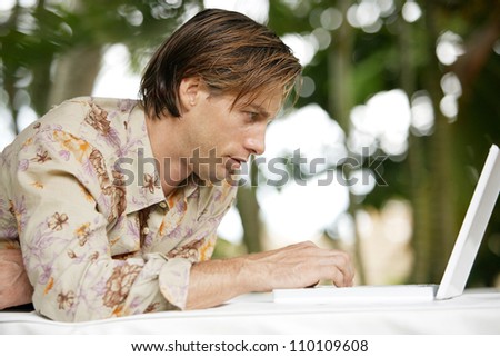 Attractive young man laying down on a outdoors bed in a tropical garden, using his laptop computer.