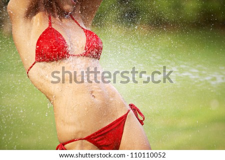 Body section of a woman wearing a red bikini in a green grass garden, having water splashed on her body in a summer day while on holiday.