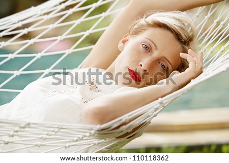 Young attractive woman laying and relaxing on a white hammock in a tropical garden near a swimming pool.