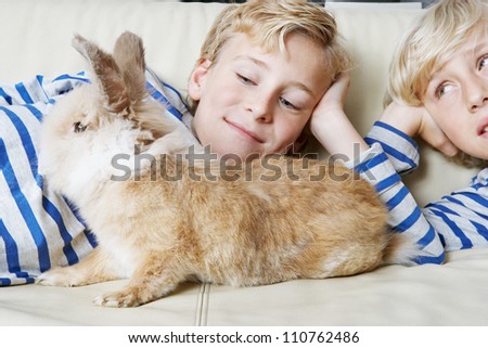 Two brothers and their rabbit pet lay together on a sofa at home, smiling.