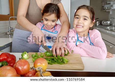 Mother and twin daughters learning to chop vegetables together in the kitchen, using a chopping board and surrounded by fruit and vegetables.