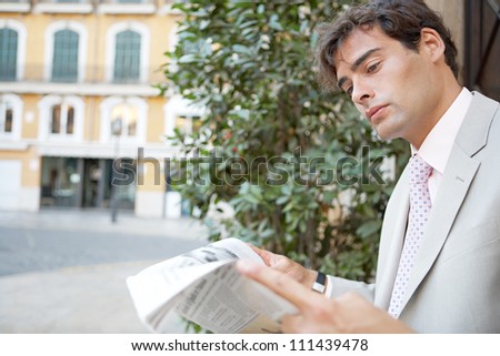 Attractive young businessman reading the newspaper while sitting in the city with classic office buildings around him.