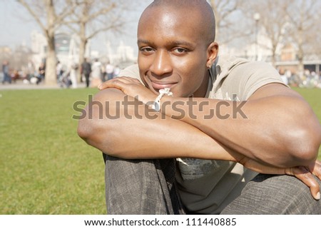 Young black tourist man sitting down on green grass in the city of London while on vacation, smiling and resting his head on his arms.