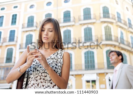 Young businesswoman using a cell phone in a classic city while a businessman walks by in the background.
