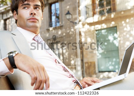 Young professional man using a laptop pc while sitting on a wooden bench in a classic city square.