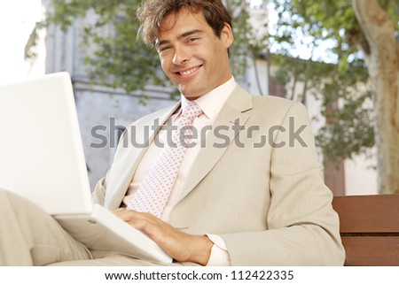 Attractive businessman sitting on a wooden bench in a classic city using a laptop computer, smiling.