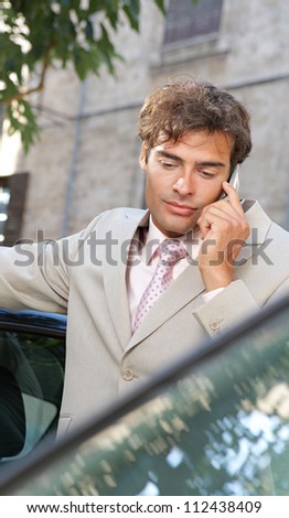Businessman using a cell phone to make a phone call while standing by some cars in the city.