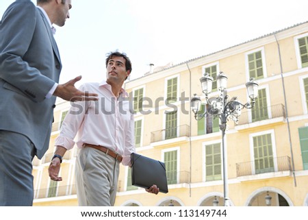 Two businessmen walking passed a building in a European city while having a conversation.