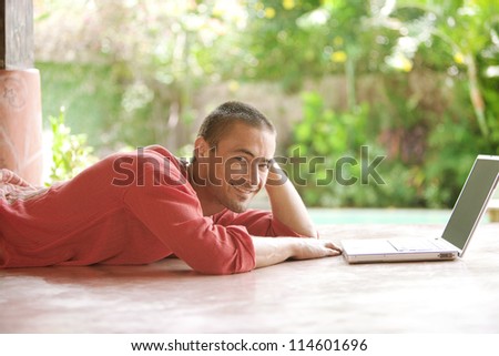 Attractive young man using a laptop computer while laying down in a tropical hotel garden, smiling.