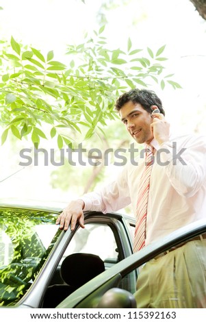 Young attractive businessman with his car having a phone conversation using a hands free set in a tree lined street in the city.