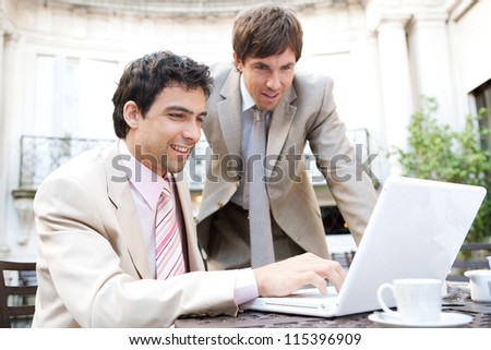Two businessmen having a meeting while sitting in a classic coffee shop terrace, using a laptop computer and smiling.