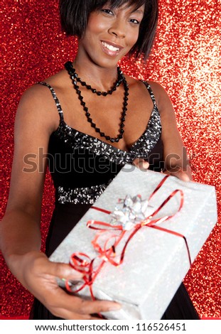 Attractive black woman holding a gift box while smiling against a red glitter background at a Christmas party.
