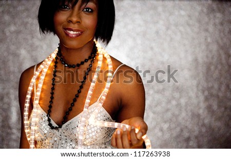 Close up portrait of a beautiful black woman holding Christmas fairy lights around her neck and smiling against a silver glitter background.