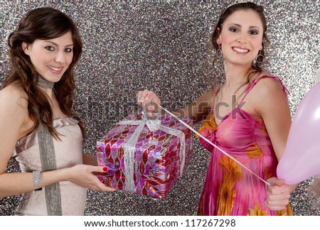Young woman offering a gift to a birthday girl at a party. Girl opening a birthday present against a silver glitter background.