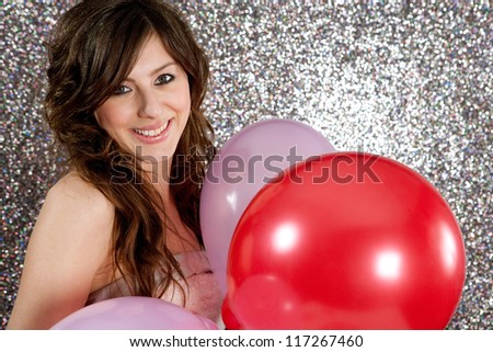 Portrait of an attractive young woman against a silver glitter background, holding three balloons in red and pink, smiling.