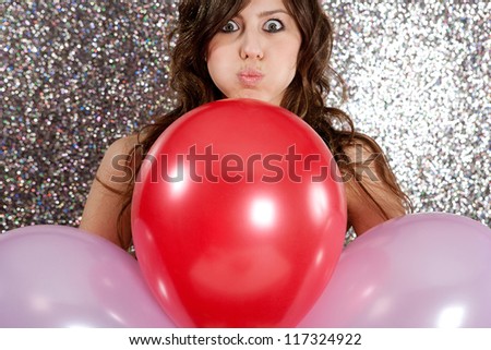 Portrait of an attractive young woman against a silver glitter background, holding three balloons in red and pink and pulling faces.