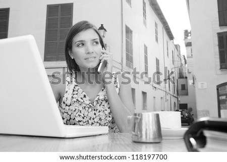 Black and white portrait of an attractive young businesswoman using a cell phone to make a call while using a laptop computer, sitting at a coffee shop terrace, outdoors.