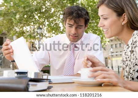 Two business people having a meeting and using technology outdoors, while having a coffee in a coffee shop terrace.