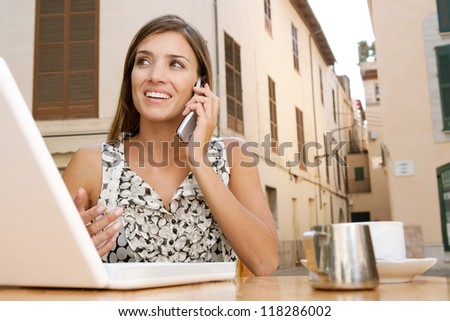 Attractive young businesswoman using a cell phone to make a call while using a laptop computer, sitting at a coffee shop terrace outdoors, smiling.